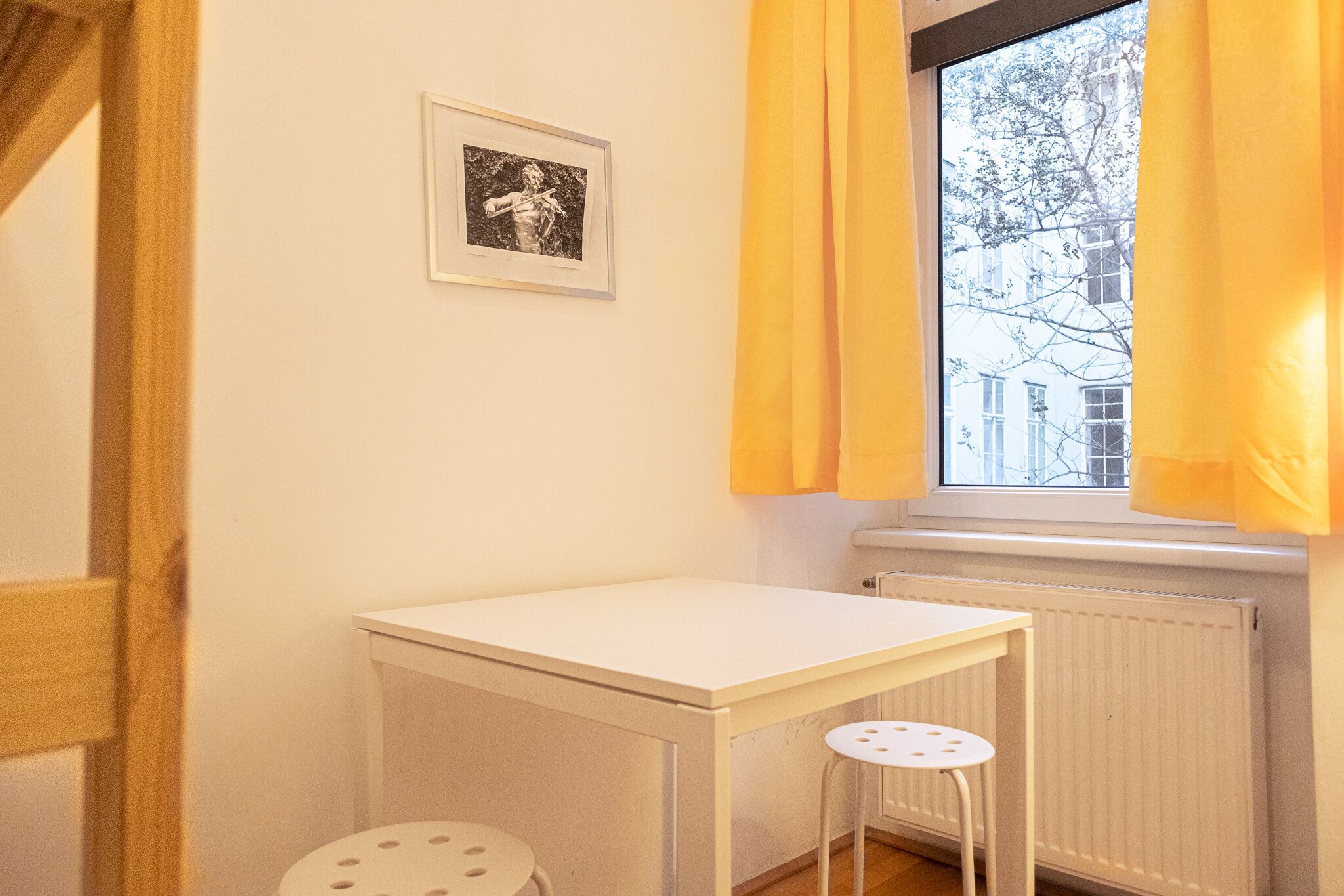 A small room with a white table, a stool, a framed picture on the wall, and a window with yellow curtains.