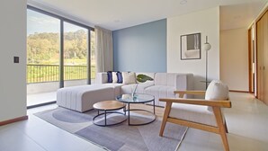 Ein modernes Wohnzimmer mit einer Eckcouch, einem kleinen runden Tisch und einem Sessel. Ein großes Fenster bietet Blick ins Grüne.