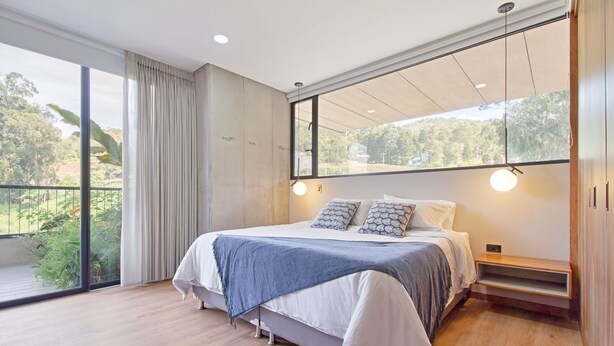 Ein modernes Schlafzimmer mit einem großen Bett, Blick ins Grüne durch raumhohe Fenster und minimalistischem Design.