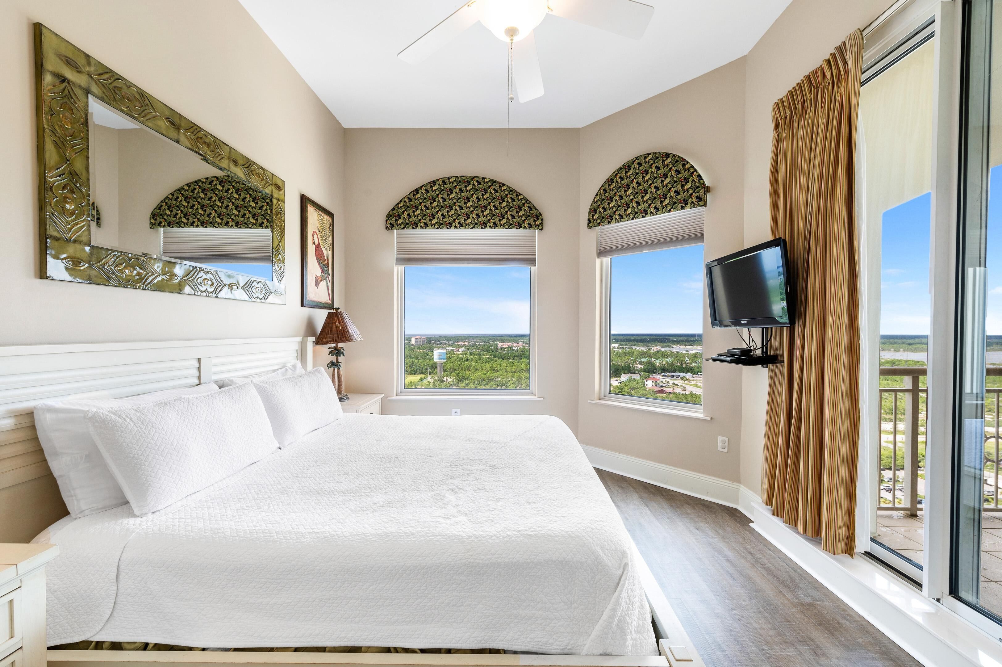 A bedroom with a large bed, a flat-screen TV, and a view of the city through large windows.
