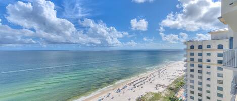 A beach with clear blue water, a sandy shore, and a hotel building with balconies overlooking the beach.