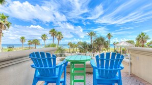 A patio with two blue Adirondack chairs and a green table, overlooking a beach with palm trees and a gazebo.