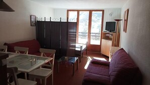 Ein kompakter Wohnbereich mit Essplatz, einer Couch, einem Fernseher und einer Schiebetür, die auf einen Balkon führt.