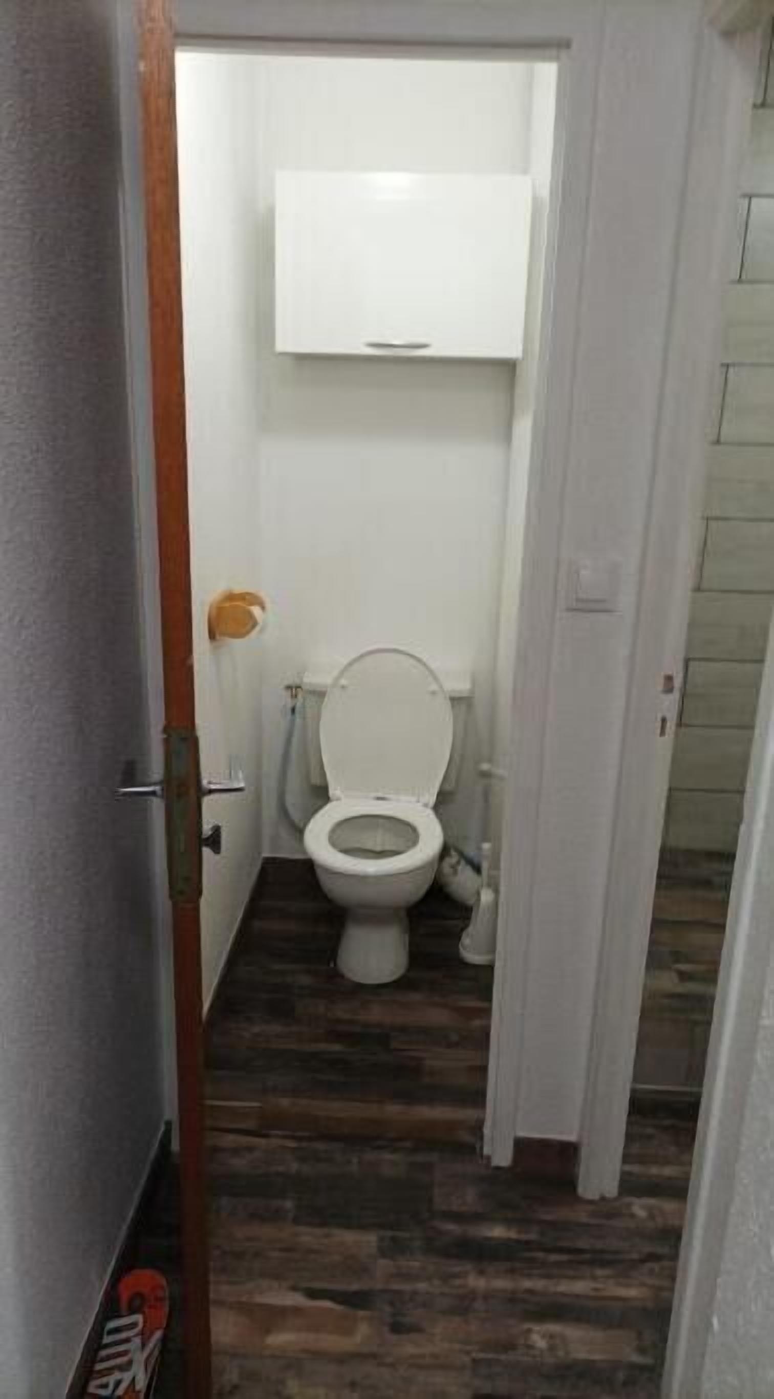 Un pasillo estrecho que conduce a un baño pequeño y funcional con inodoro, un armario blanco encima y piso de madera.