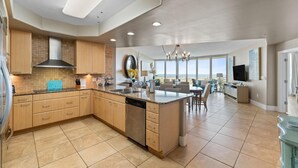 A spacious kitchen with a large island, wooden cabinets, and a dishwasher. There is a dining area with a table and chairs, and a view of the ocean through large windows. - Caribe D411 (Orange Beach)