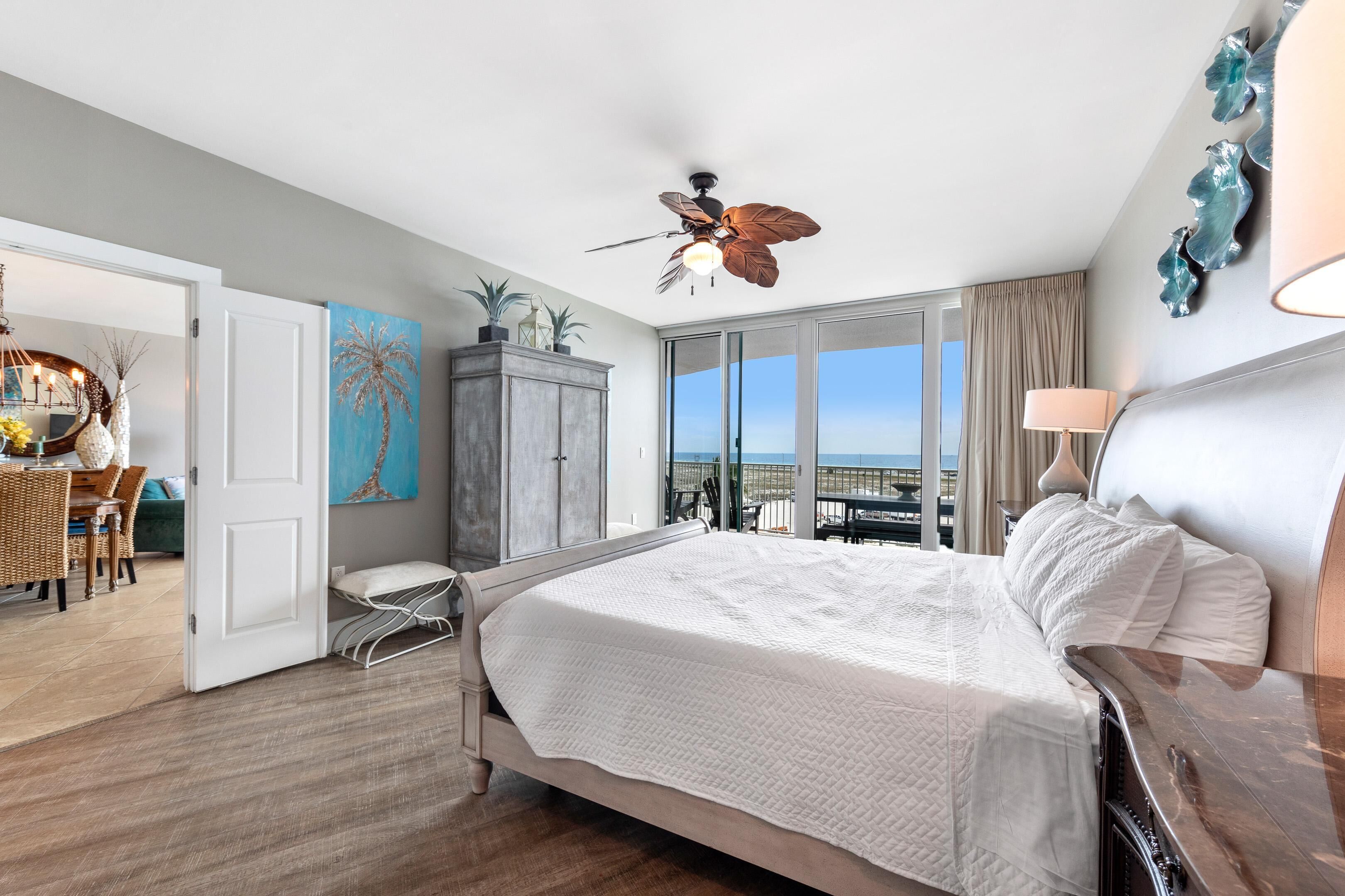 A hotel room with a large bed, a ceiling fan, a view of the ocean, and a dining area with wicker furniture.