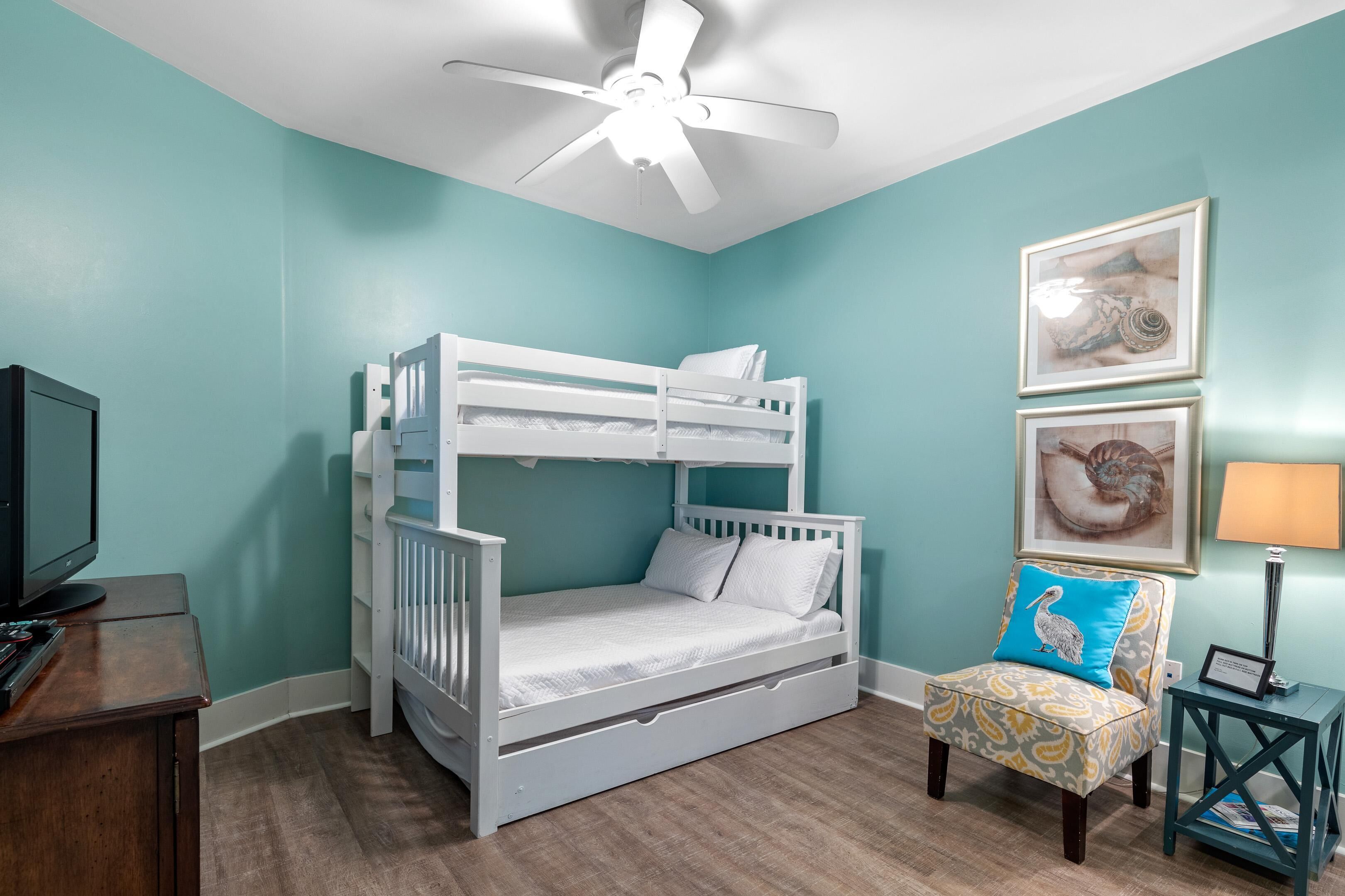 A room with a bunk bed, a desk with a computer, a chair with a patterned cushion, and a ceiling fan.