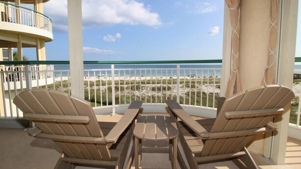 Un balcon avec deux fauteuils Adirondack et une table, donnant sur une plage délimitée par une clôture, sous un ciel dégagé.