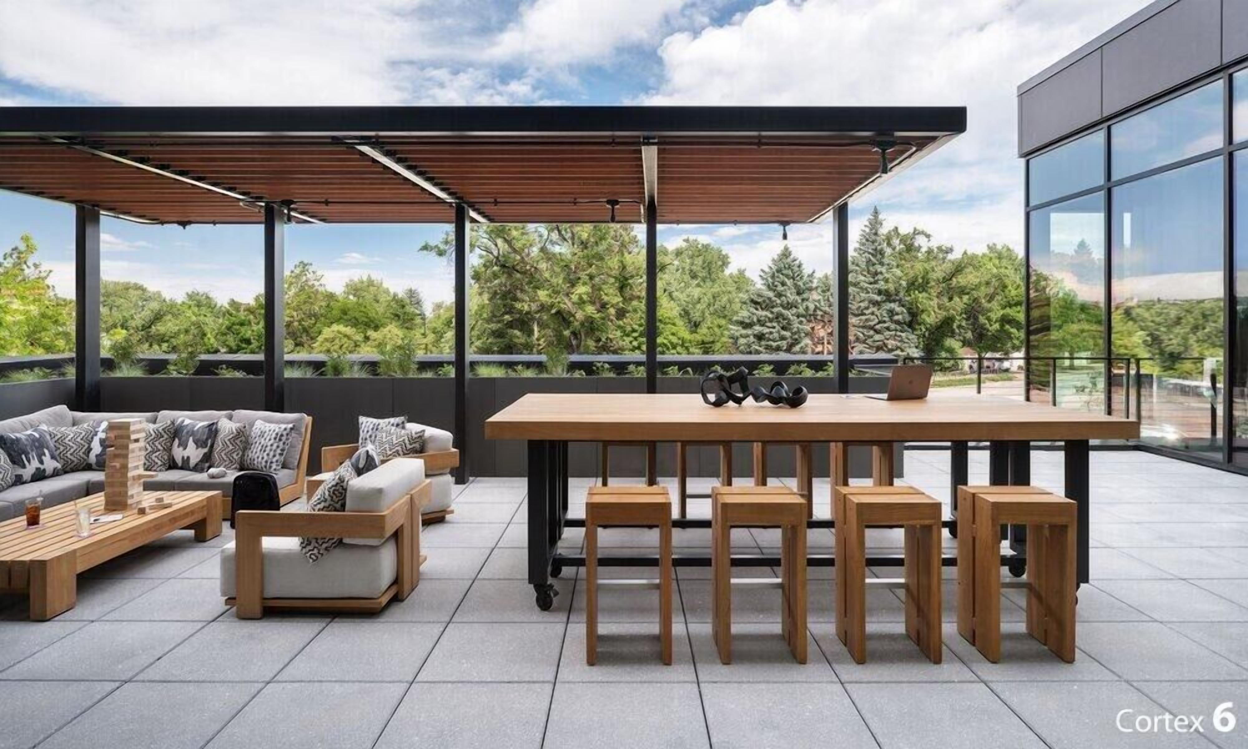 Un espacio moderno al aire libre con una mesa de madera y bancos, un sofá, y una computadora portátil sobre la mesa.