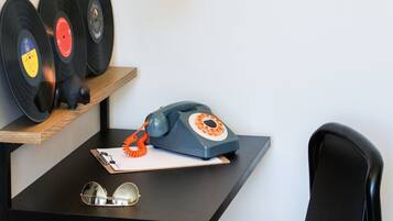A room with a desk, a chair, a lamp, a shelf with vinyl records, a rotary phone, and a framed picture on the wall.