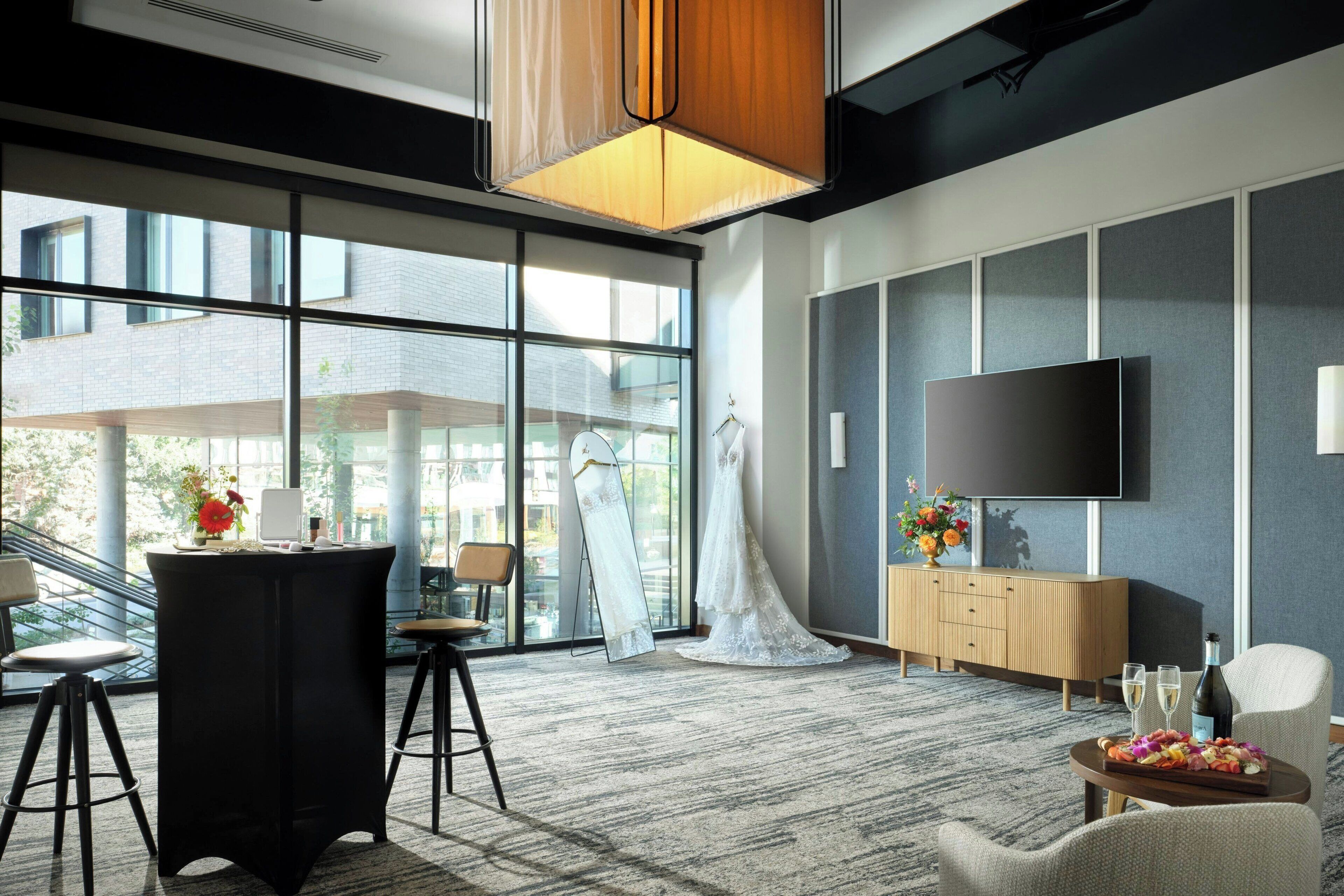 A modern hotel lobby with a large window, a flat-screen TV, a wooden cabinet, and a round table with a fruit platter.