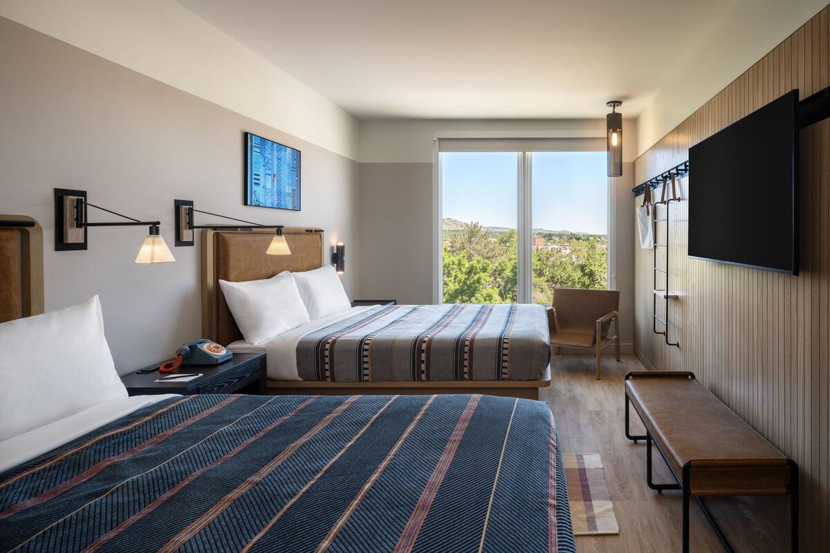 Habitación de hotel con dos camas, un escritorio, una silla, un televisor y un amplio ventanal con vistas al exterior.