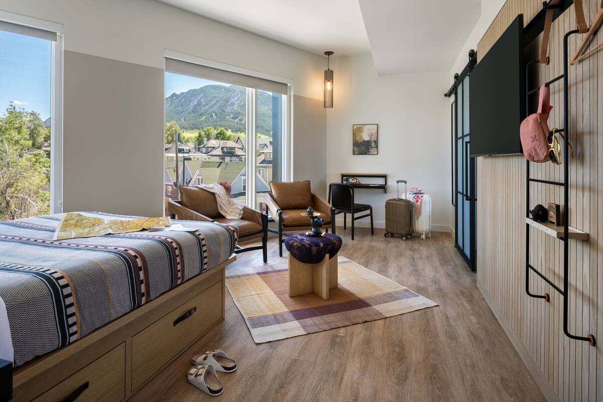 A modern bedroom with a large bed, a chair, a desk, and a view of the mountains.