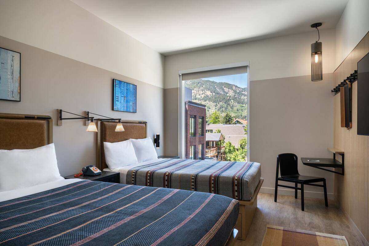 A hotel room with two beds, a desk, a chair, a large window with a mountain view, and artwork on the walls.