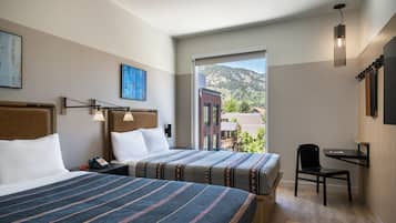 Quarto de hotel com duas camas, uma escrivaninha, uma cadeira, uma janela ampla com vista para a montanha e obras de arte nas paredes.