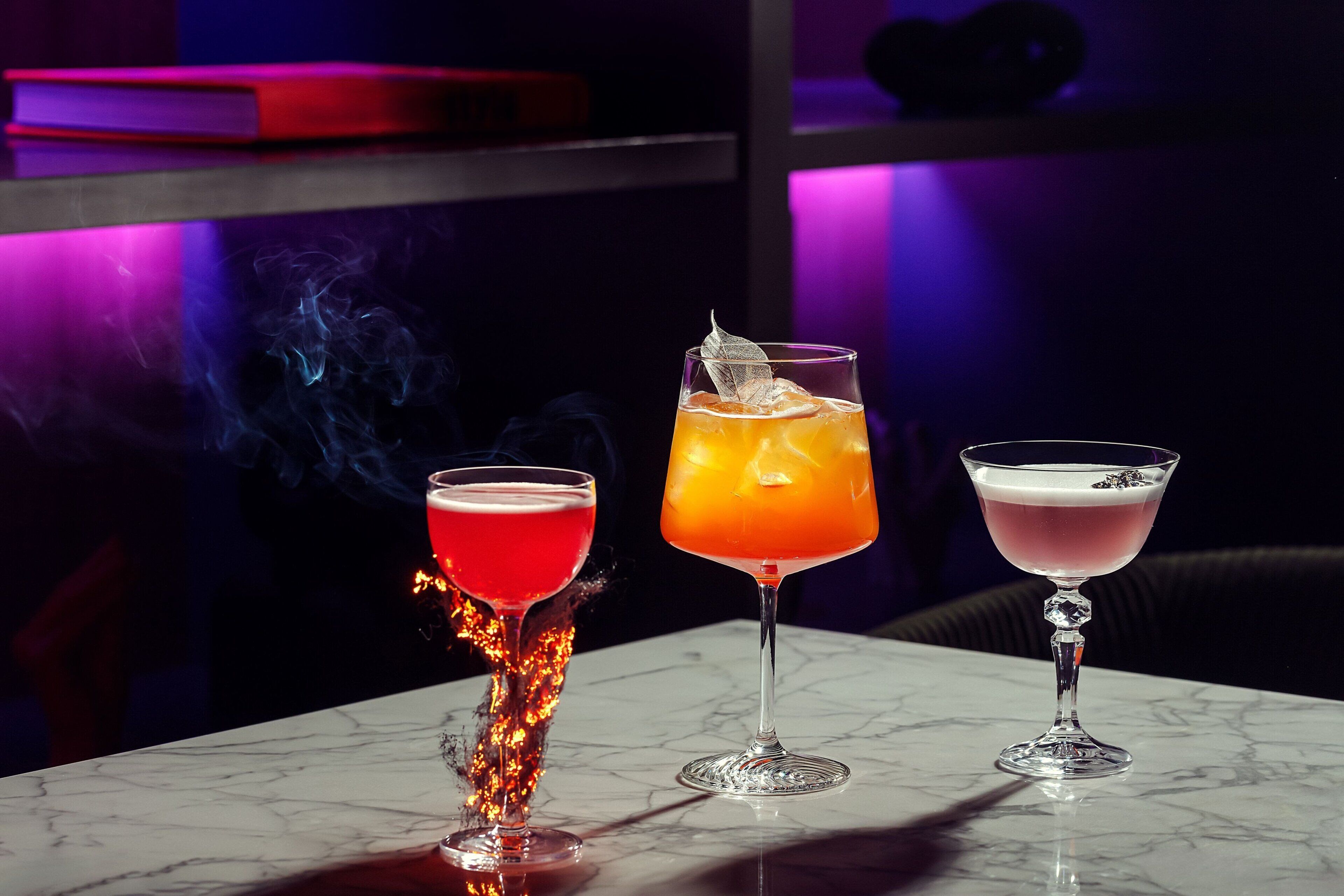 Drei Cocktails auf einem Marmortisch, vor dunklem Hintergrund und violetter Beleuchtung.