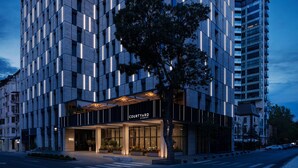 Un hotel moderno con fachada geométrica, un acceso por patio interior y un árbol en primer plano.