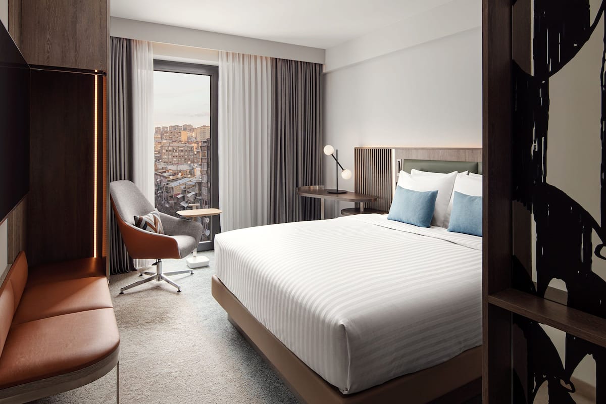 A hotel room with a large bed, a seating area with a chair, a small table with a lamp, and a view of the city through the window.