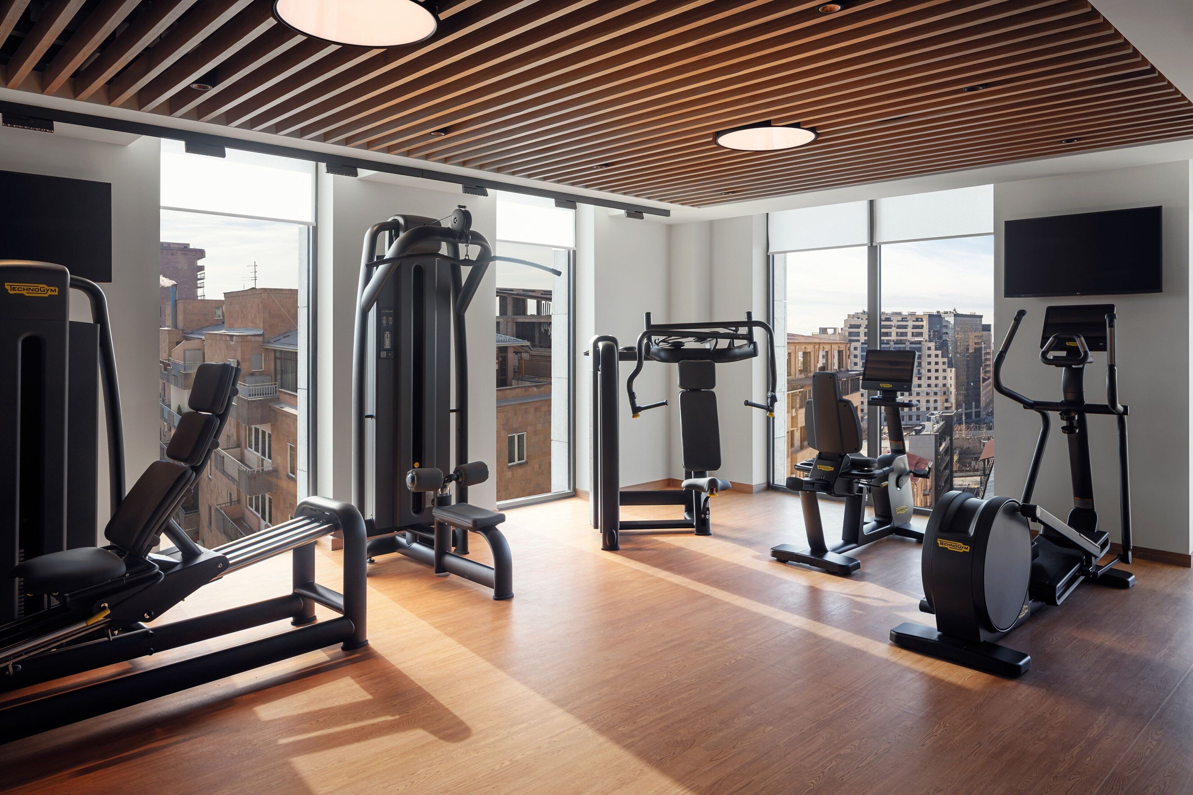 A gym with various exercise machines, large windows, and a wooden ceiling.