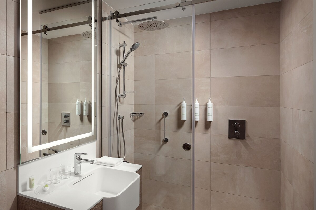 Un baño moderno con ducha de vidrio, un espejo grande y un lavabo blanco con artículos de aseo.