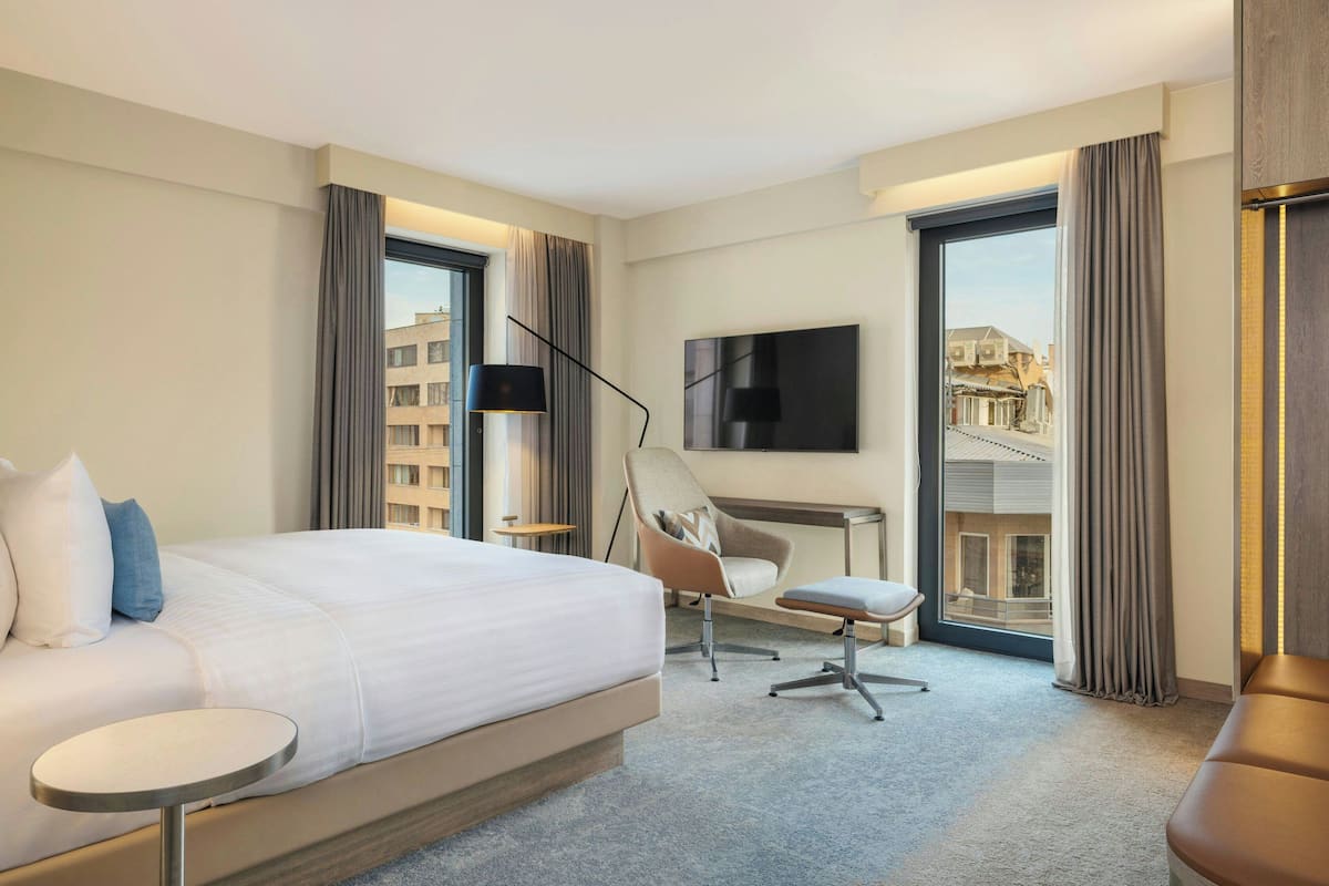 A hotel room with a large bed, a desk, a chair, a TV, and a view of buildings through the window.
