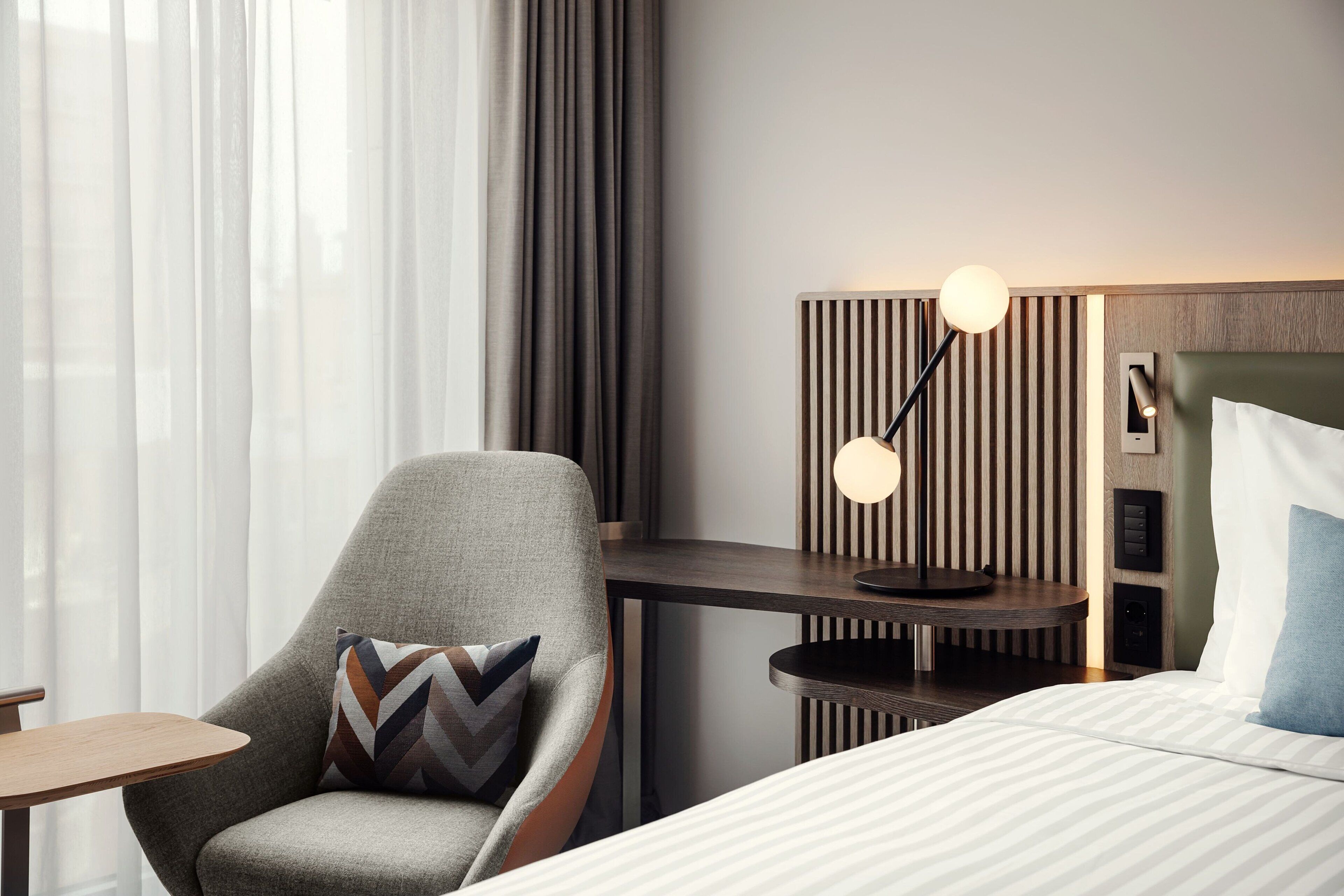 A hotel room with a bed, a chair with a patterned pillow, a bedside table, a lamp, and a wall-mounted control panel.