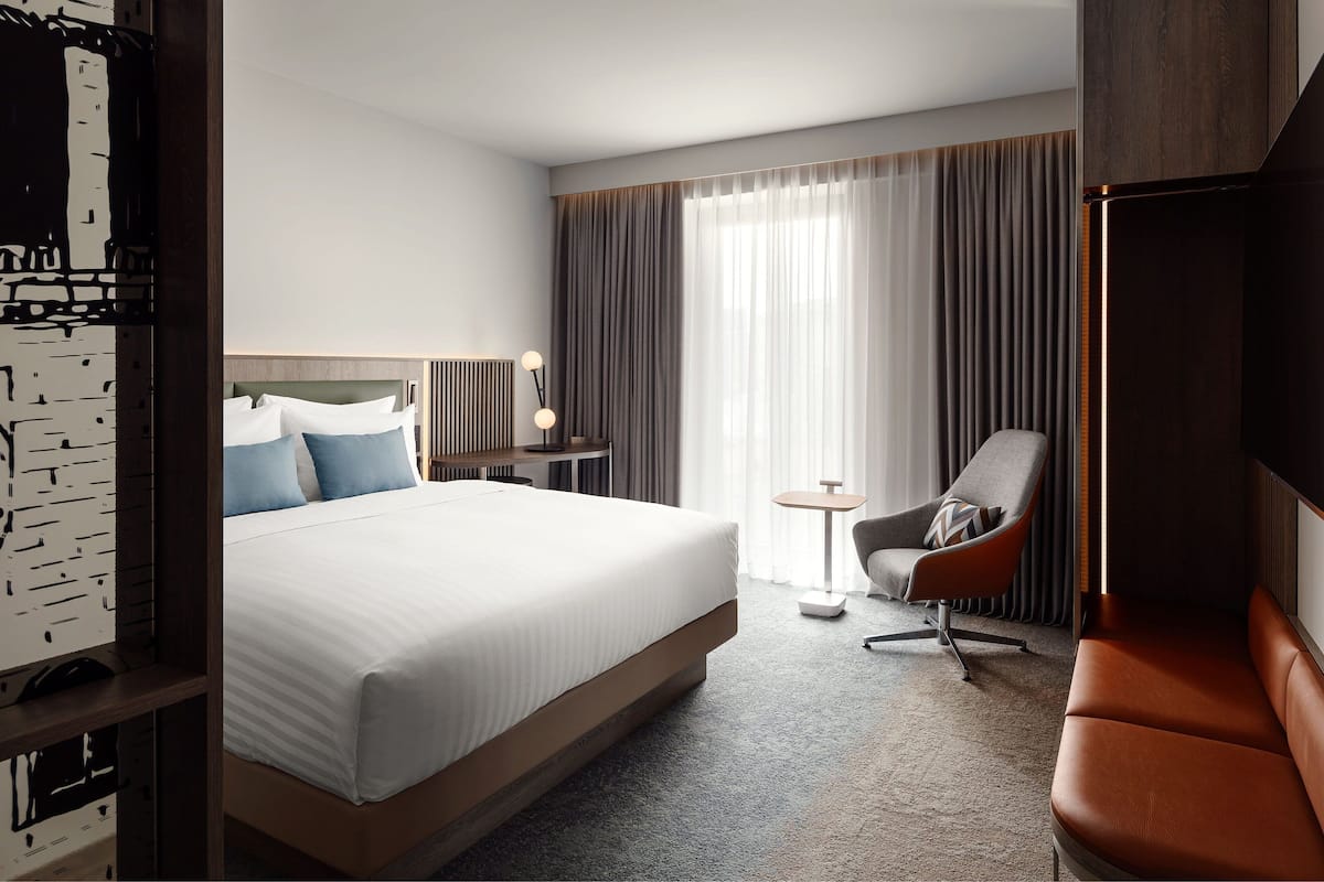 A hotel room with a large bed, a chair, a small table, and a window with curtains.