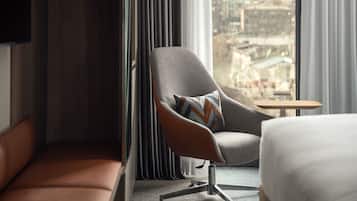 A hotel room with a modern armchair, a small table, a bed, and a view of the city through large windows.