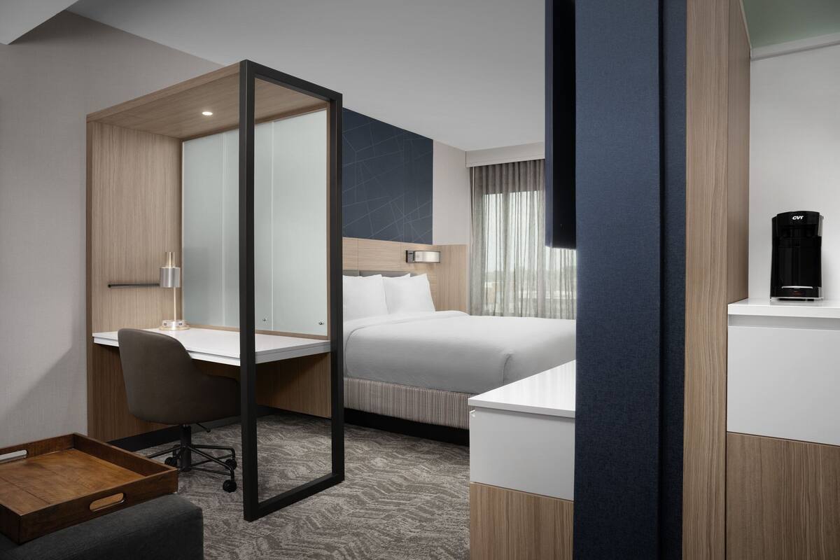 A modern hotel room with a bed, a desk, a chair, a mirror, and a coffee maker.
