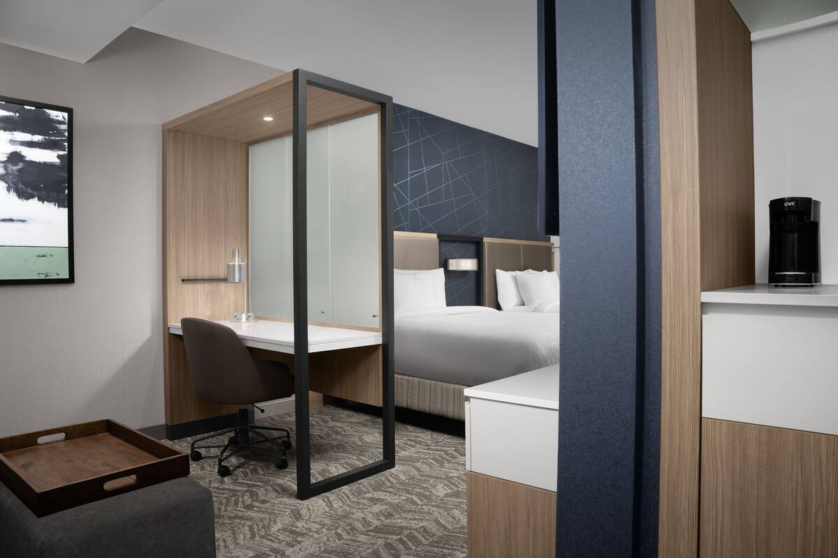 A modern hotel room with a bed, desk, and chair, a coffee maker, and a framed picture on the wall.