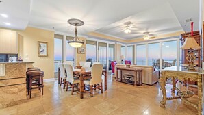 A spacious living area with a dining set, a large table, and a view of the ocean through large windows. - La Playa 1001 (Pensacola)