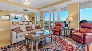 A spacious living room with a large sofa, leather armchairs, a glass coffee table, and a view of the outdoors through large windows. - La Playa 1001 (Pensacola)