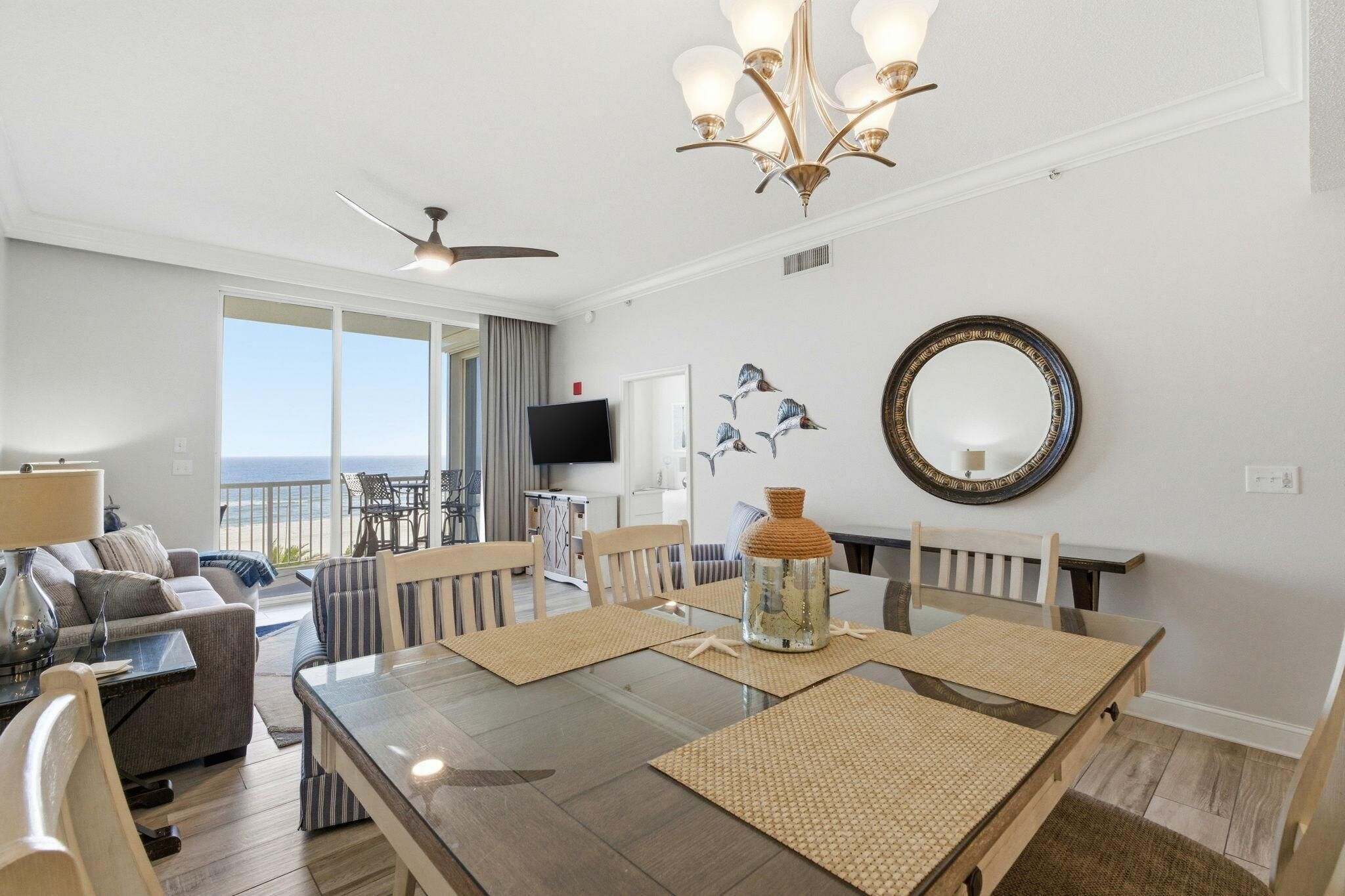 A modern living room with a glass dining table, comfortable seating, a ceiling fan, and a large window overlooking the ocean.