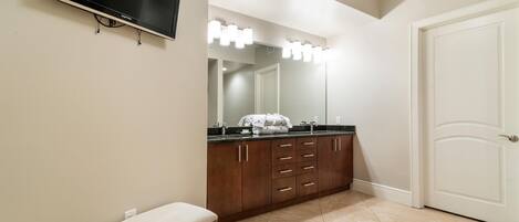 Un baño con tocador, espejo con iluminación, televisor y puerta.