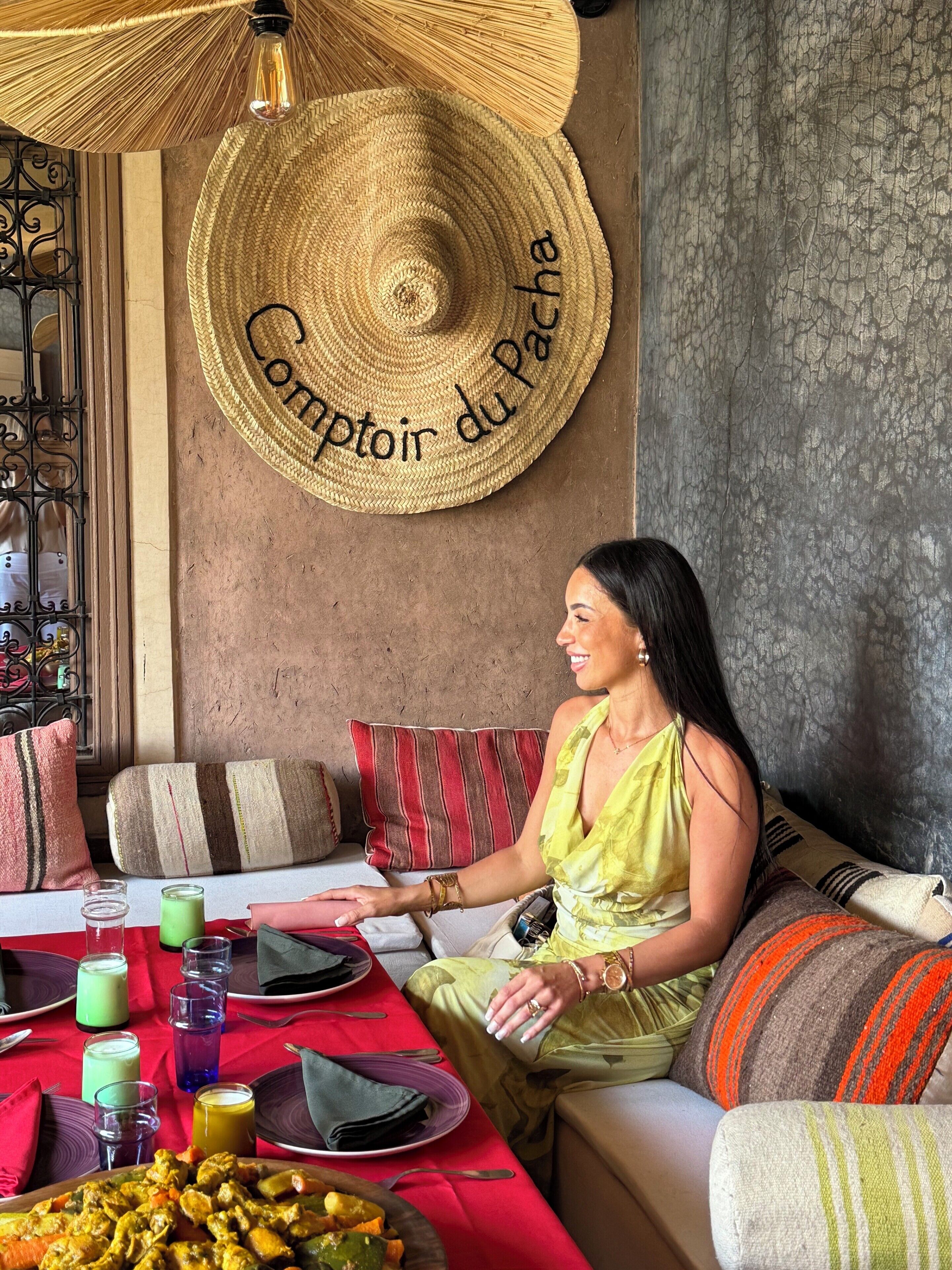 A person seated at a table with a variety of dishes, a large woven hat on the wall, and a textured wall behind.