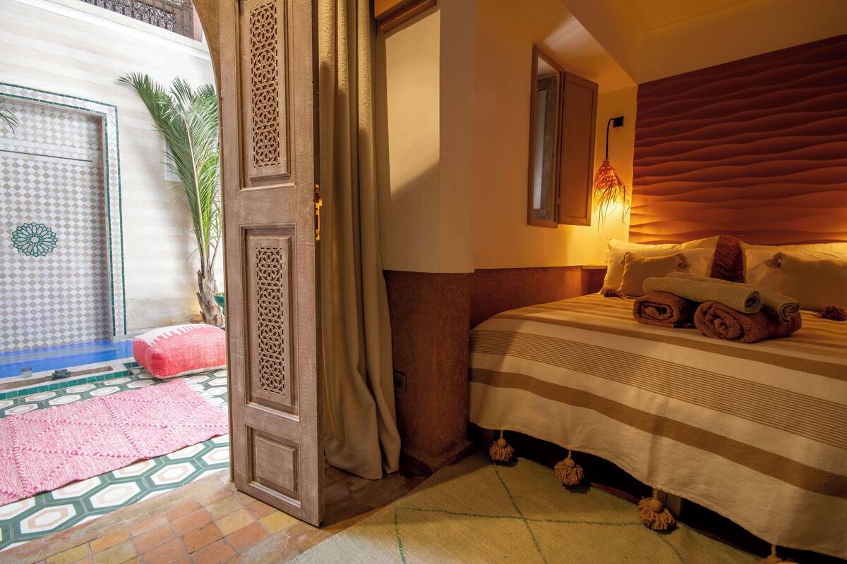 A bedroom with a bed, pillows, a nightstand, a lamp, and a door leading to a balcony with a view.