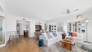 A spacious living room with a white sofa, a wooden coffee table, and a large rug. There is a dining area with a table and chairs, and a kitchen in the background. - Indigo West 1101 (Pensacola)