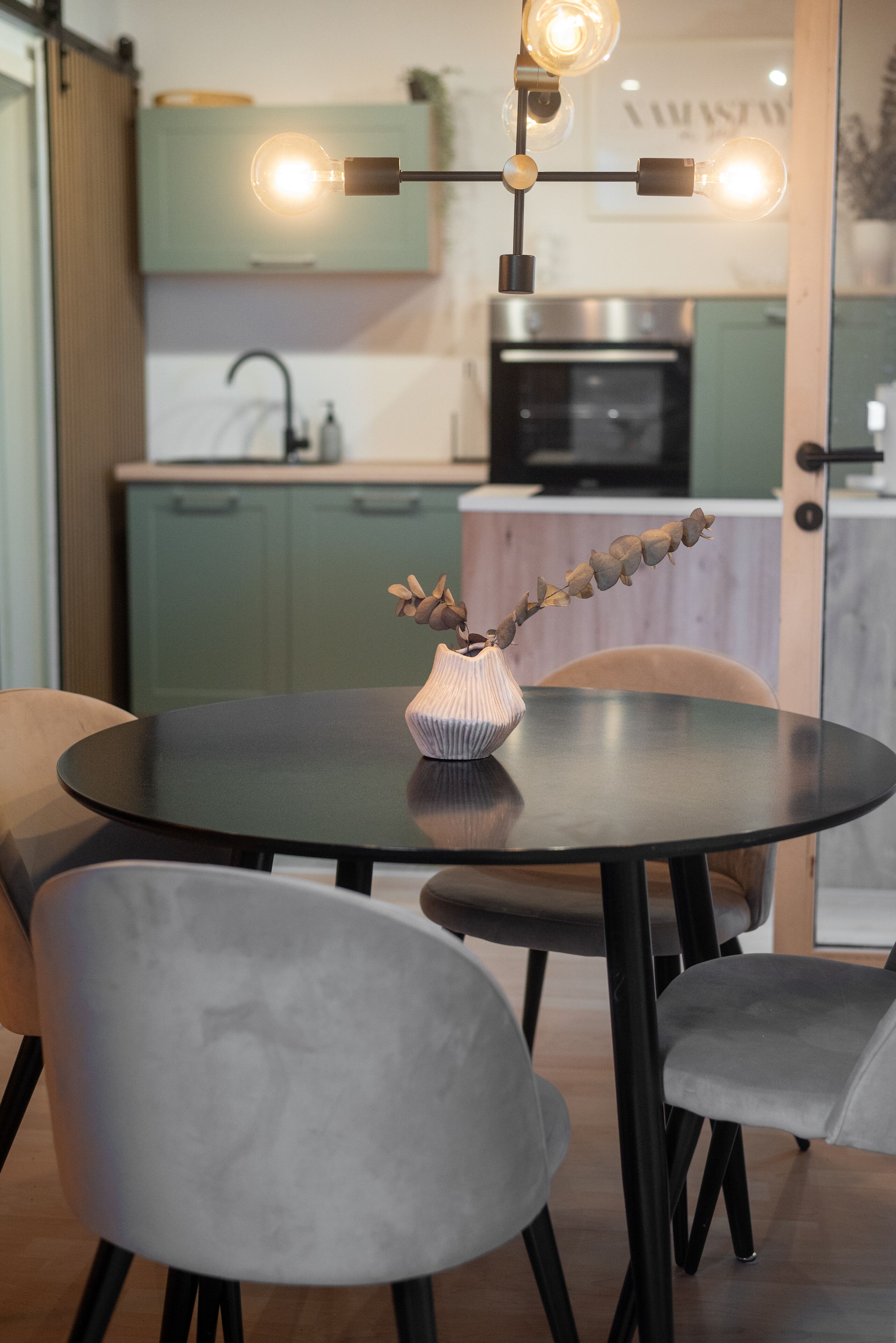 Un coin repas avec une table ronde, quatre chaises, un vase avec des fleurs séchées et, au fond, une cuisine.