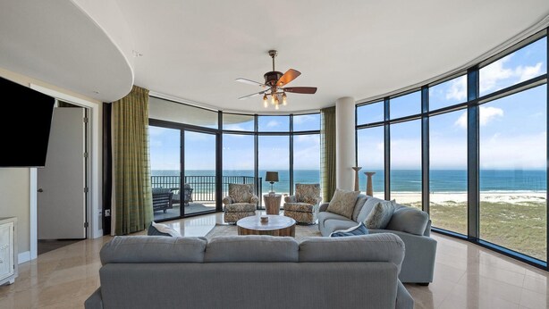 A spacious living room with a large gray sofa, a round coffee table, and a ceiling fan. The room features large windows offering a view of the beach and ocean. - Capri 302 (Pensacola)