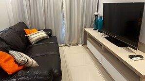 A living room with a black leather sofa, a flat-screen TV, and a white console table. - Brisas do Lago - Apartamento 12 (Brasília)