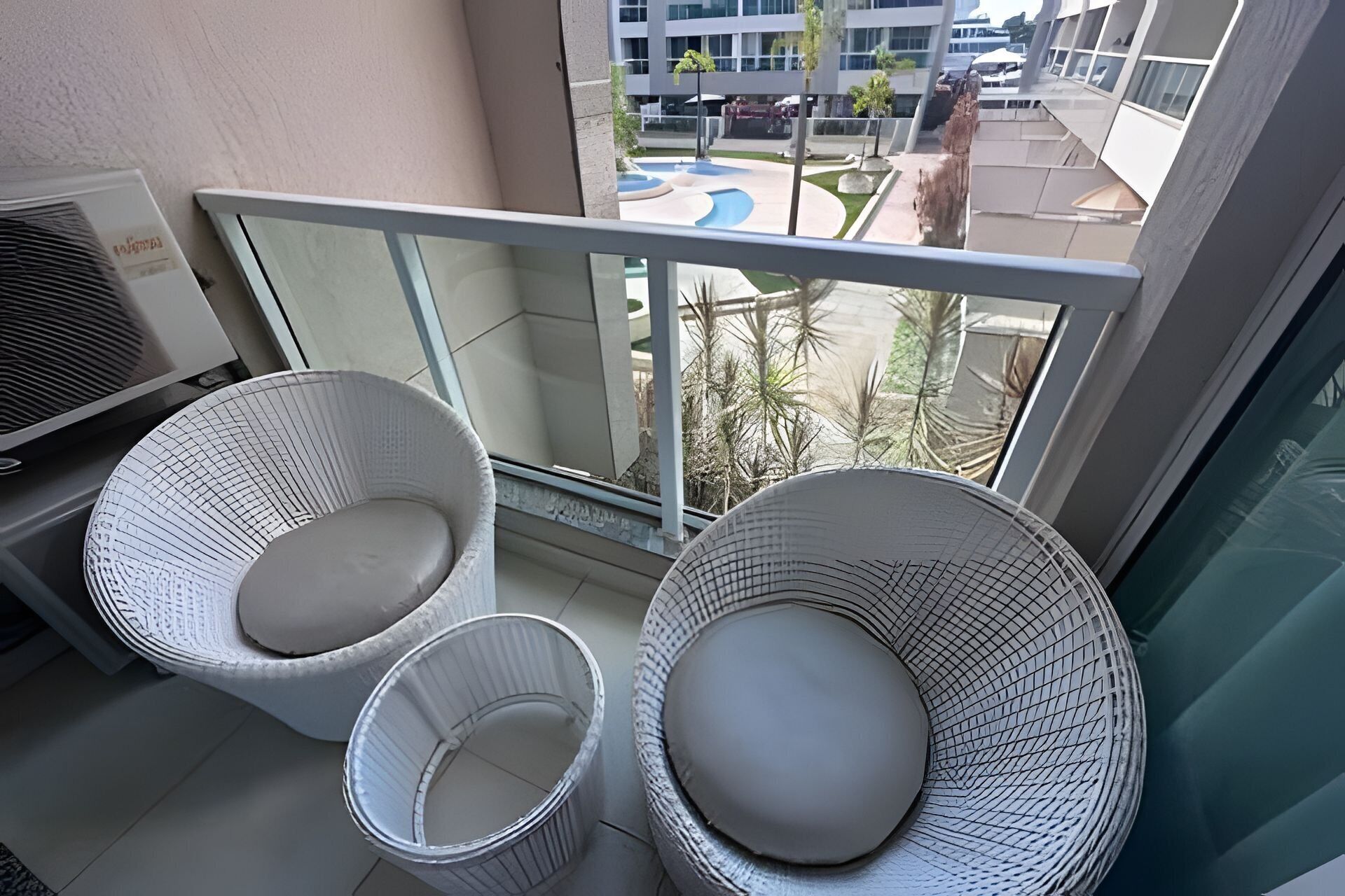 Un balcón con dos sillas de mimbre y una pequeña mesa, con vistas a una piscina y edificios.