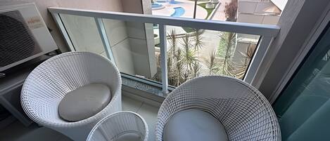 Un balcón con dos sillas de mimbre y una pequeña mesa, con vistas a una piscina y edificios.
