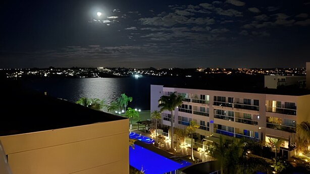 A nighttime view of a coastal cityscape with a prominent building featuring multiple floors and balconies, a body of water in the foreground, and a visible moon in the sky. - Brisas do Lago - Apartamento 12 (Brasília)
