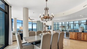 A spacious dining area with a large table and comfortable chairs, a chandelier, and a view of the ocean through large windows. - Capri 601 (Pensacola)