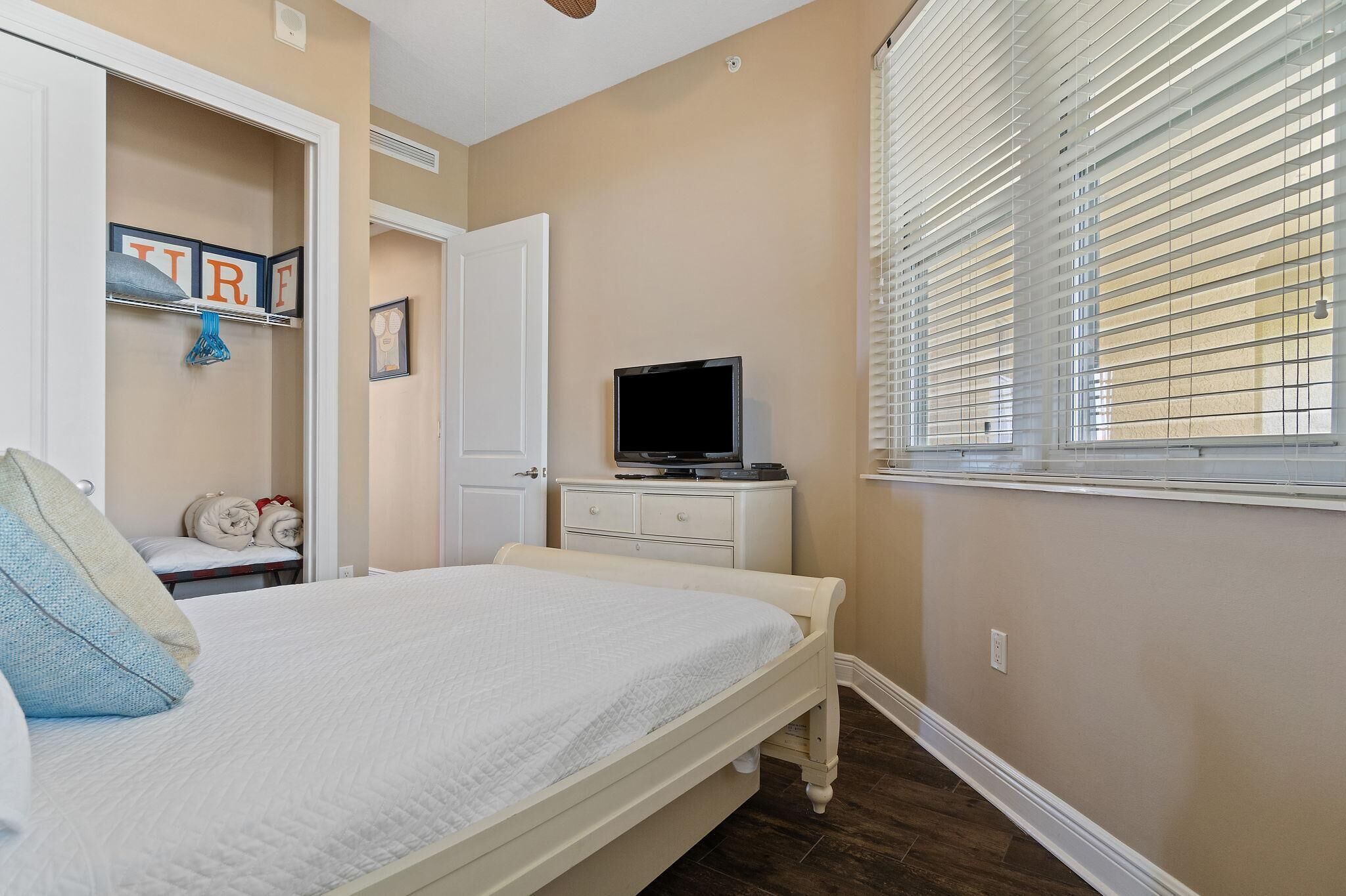 A bedroom with a bed, a TV on a dresser, a window with blinds, and a door leading to another room.