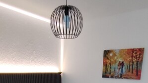 Un lit bien fait, avec une couette blanche, un coussin décoratif et une serviette en forme de cygne. Une lampe suspendue moderne est accrochée au-dessus du lit, et un tableau coloré orne le mur.