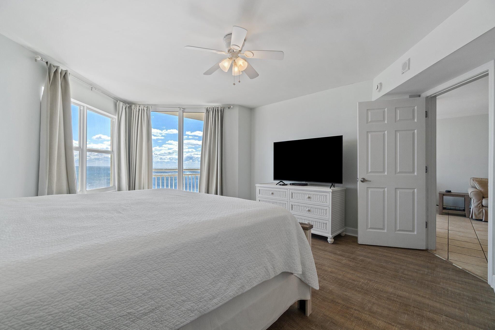A bedroom with a large bed, a ceiling fan, a TV, and a door leading to another room.