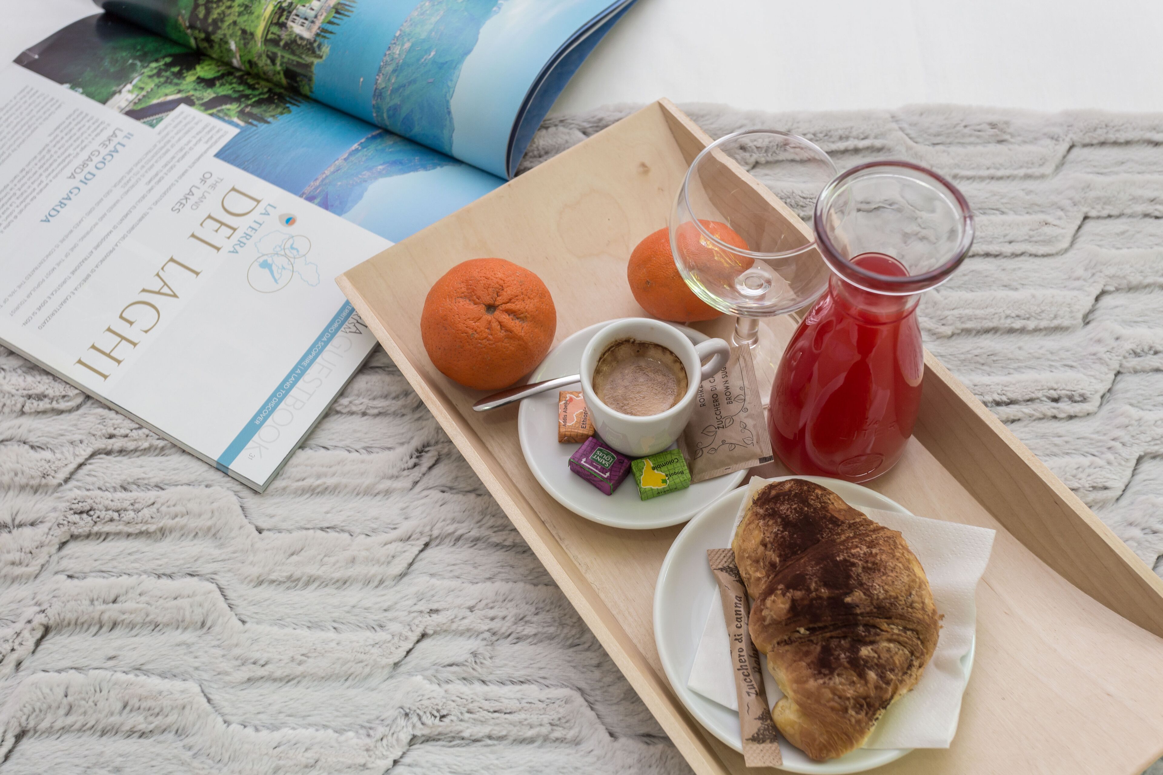 A tray with a croissant, orange, coffee, and a glass of red drink on a textured surface with a book and magazine.
