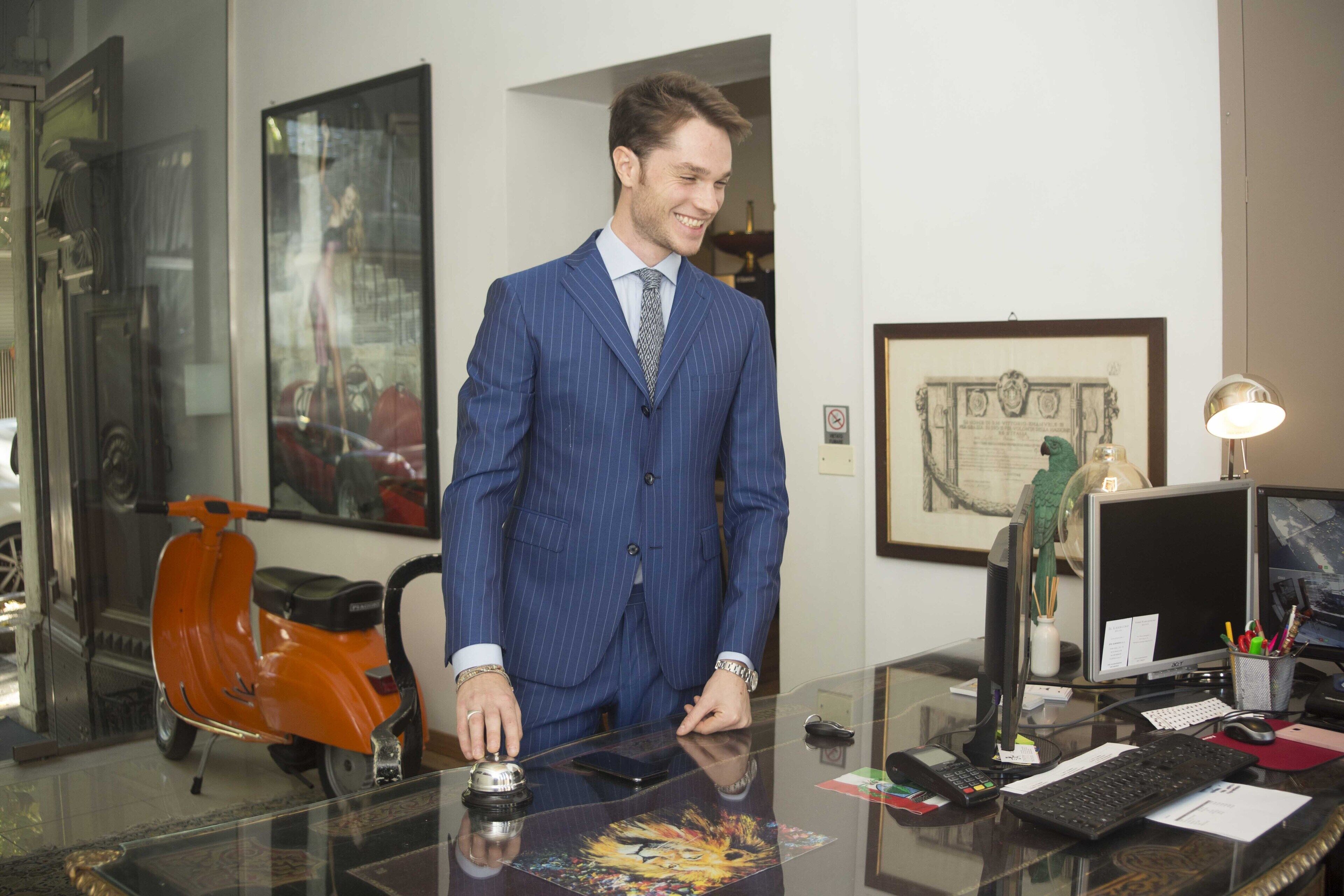A man in a pinstripe suit standing in a room with a desk, a computer, and an orange scooter.