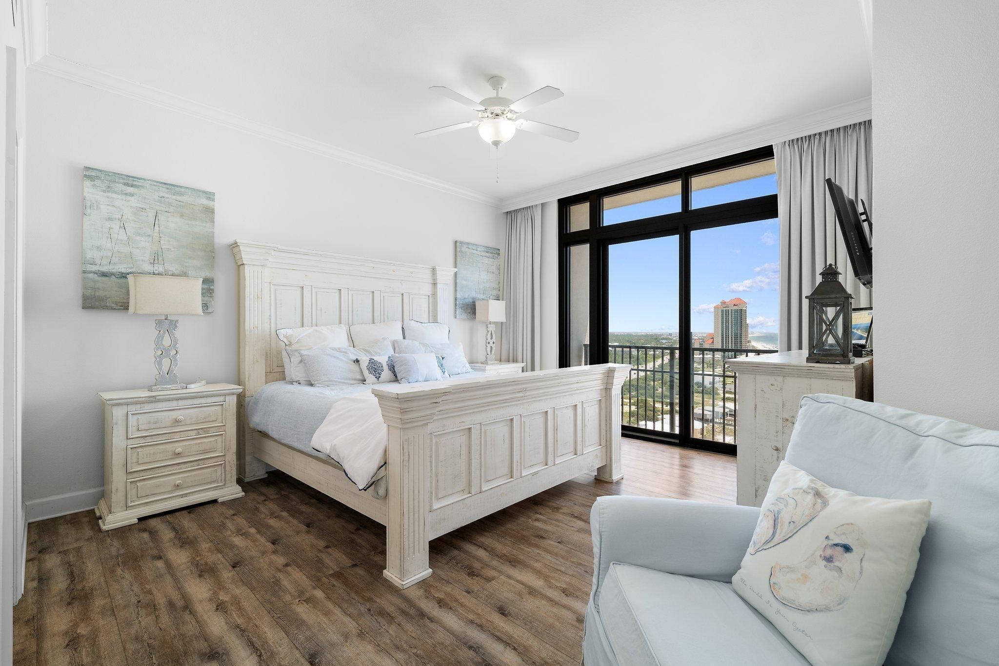A bedroom with a large bed, a nightstand, a lamp, a ceiling fan, and a view of the city through large windows.